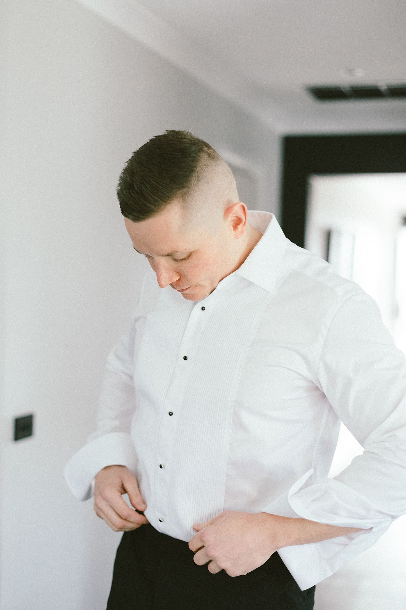 A person in a crisp white dress shirt buttons up while getting ready in a bright room.