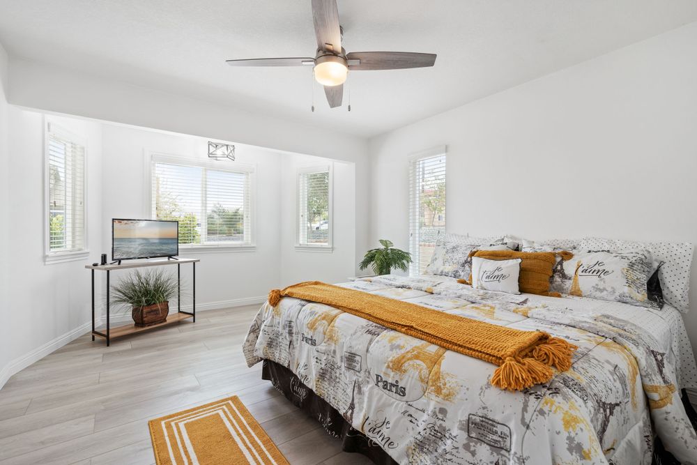 Bright bedroom with gray and yellow bedding, hardwood floors, and large windows.