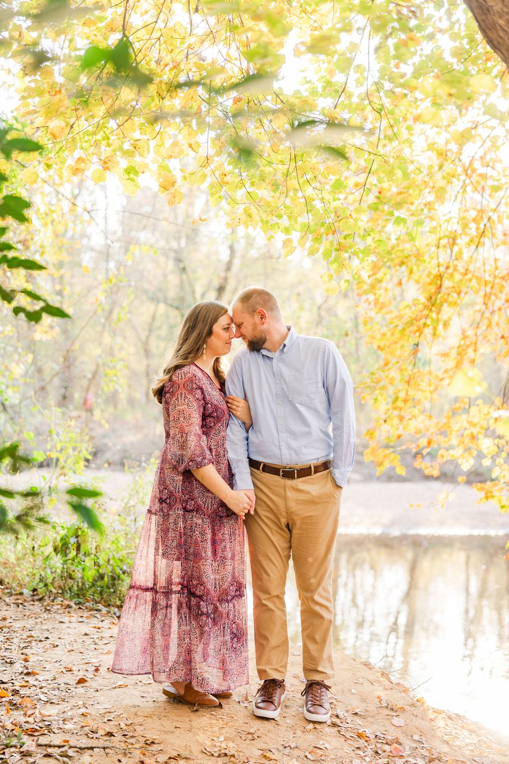 A romantic autumn photo shoot along a tree-lined path by a river with warm golden foliage overhead.