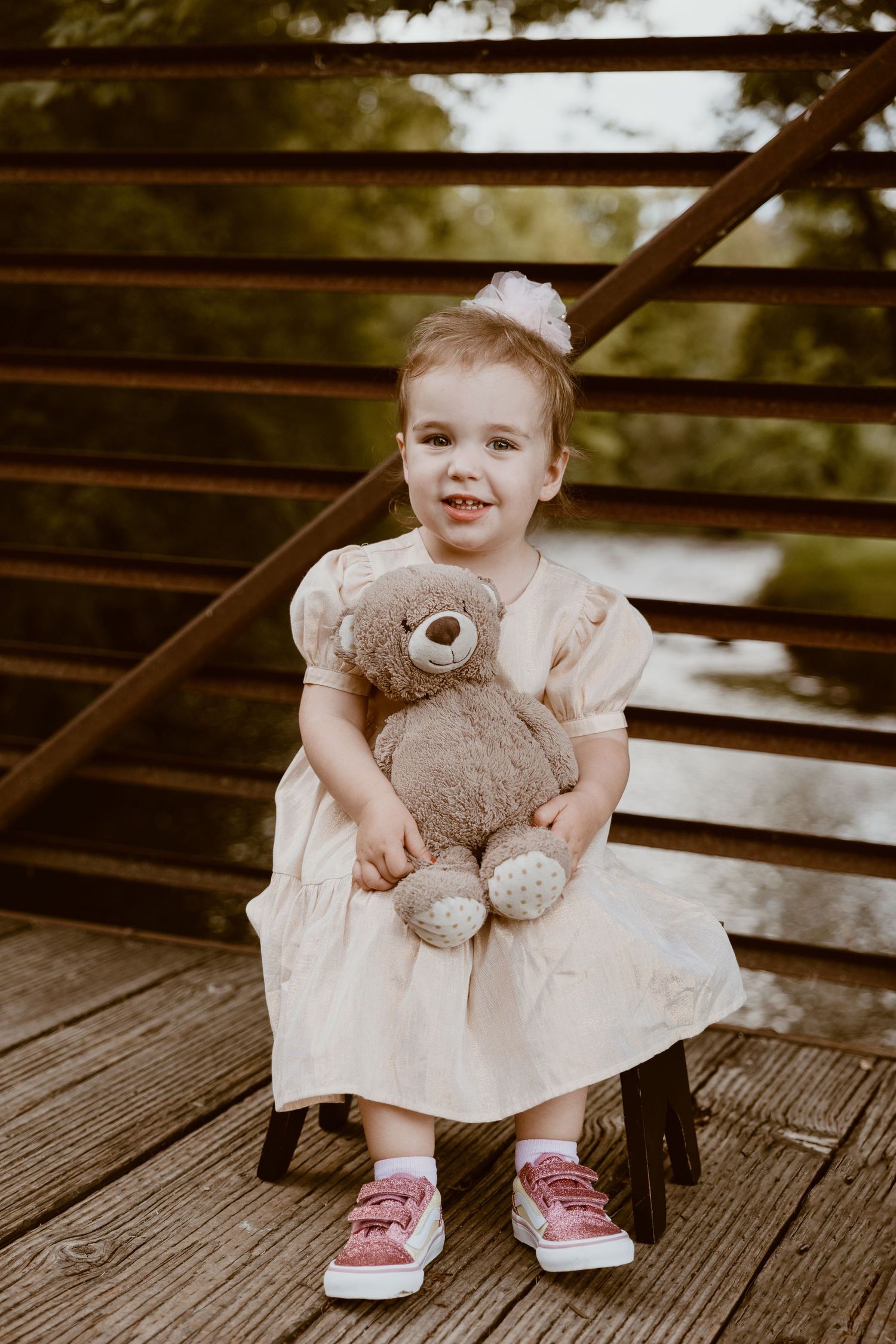 A child wearing a dress and red sneakers sits on wooden stairs while holding a plush teddy bear.