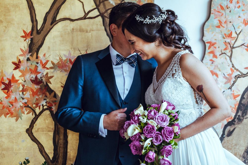 Bride and groom share intimate moment against cherry blossom mural with purple bouquet.
