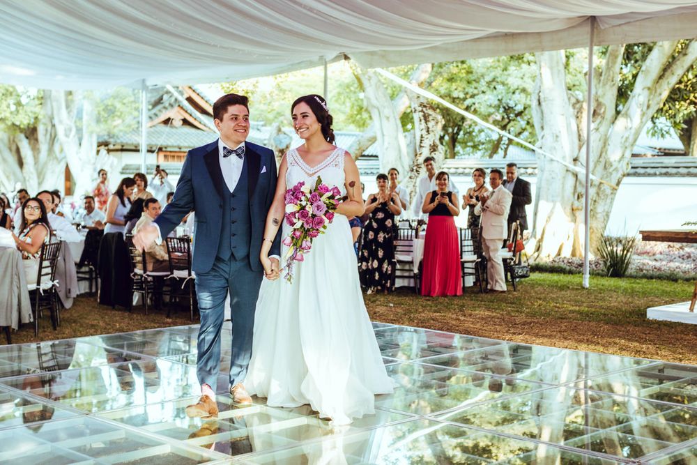 Newlyweds walking on reflective floor under white tent with guests in background.