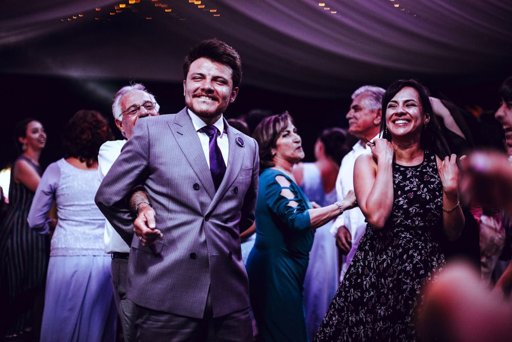 Wedding guests dancing and celebrating on a lively reception dance floor under purple lighting.