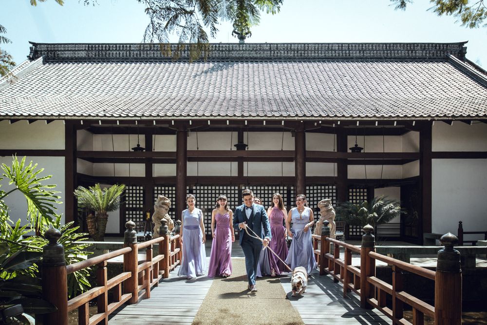 Wedding party poses on wooden bridge walkway in front of traditional Japanese-style building.