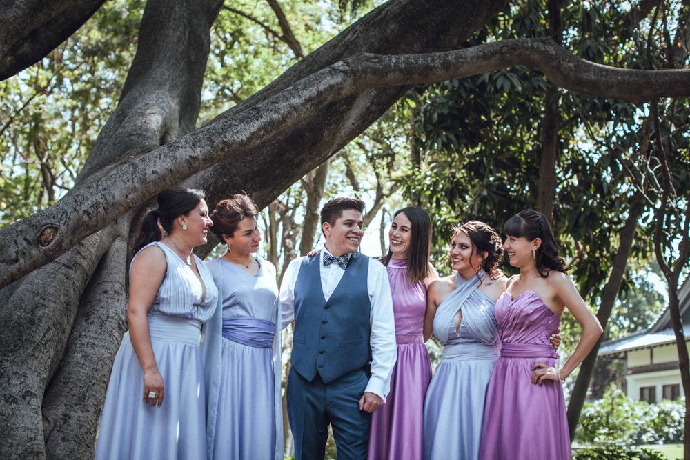 Wedding party poses together under large tree branches in purple and grey formal attire.