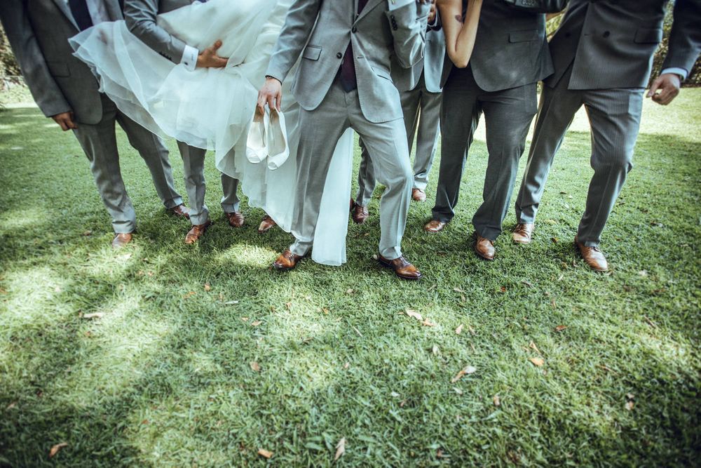 Wedding party's feet and legs gathered on grass showing formal shoes and dress hem.