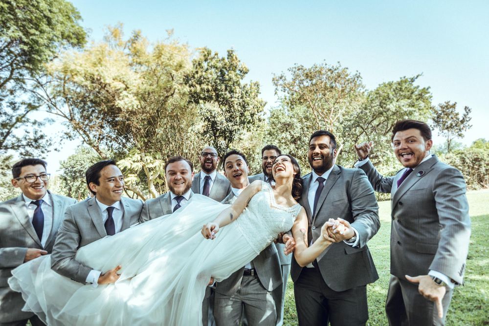 Wedding party in grey suits and light blue dresses playfully lifting veils outdoors at a sunny celebration.