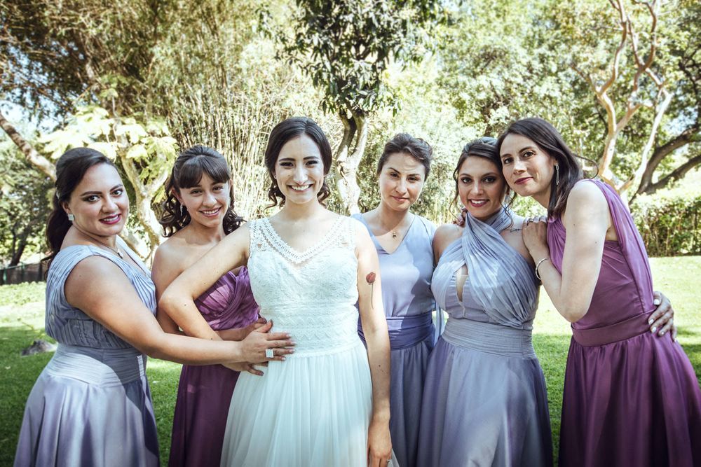 Bridesmaids in purple and blue dresses pose with bride in white gown on grassy lawn.