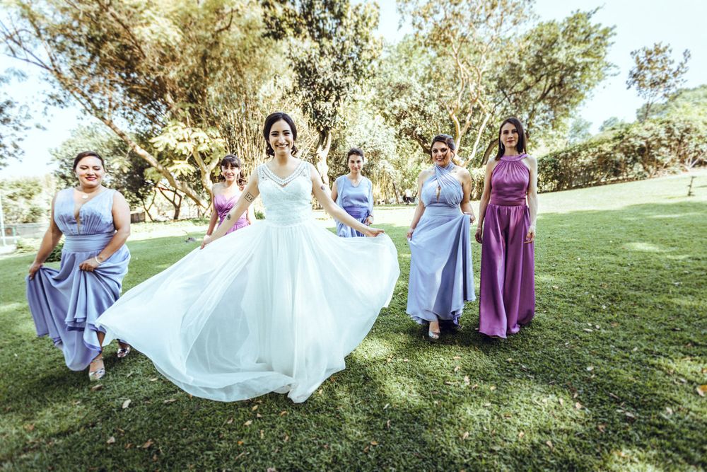Bridesmaids help adjust bride's dress while sitting on grass during outdoor photo session.