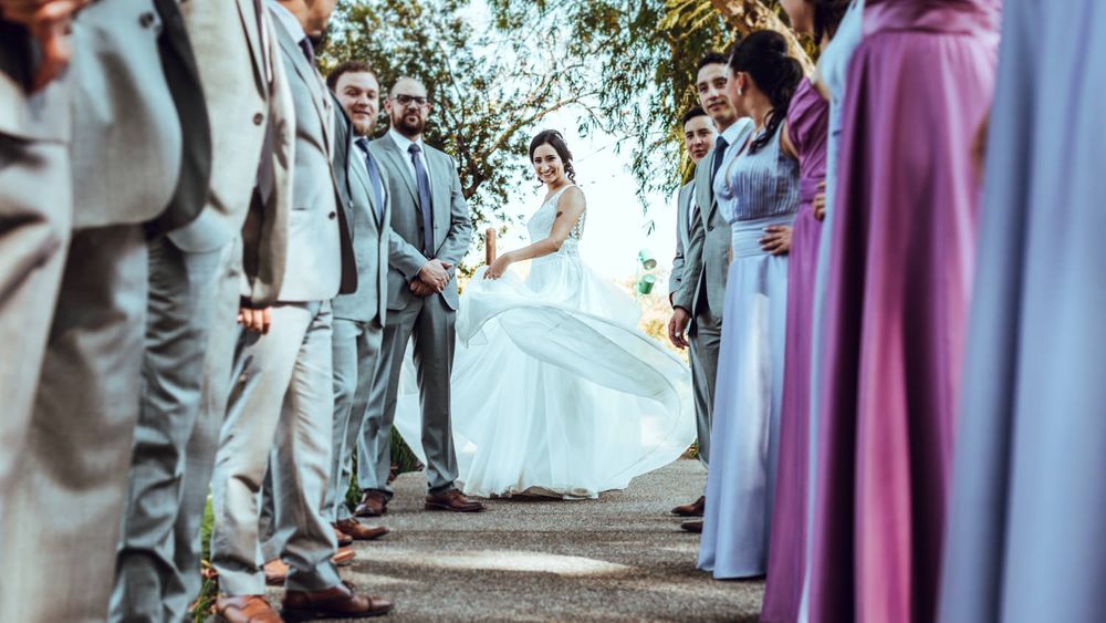 Wedding party in purple dresses and grey suits pose together under trees.