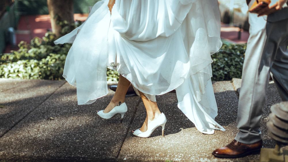 Close-up of bride's white dress hem and shoes walking on garden path.