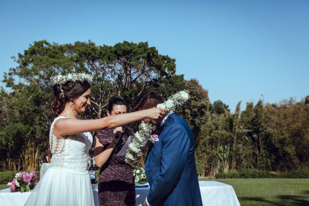 A newlywed couple playfully shares a moment with floral decorations during their outdoor celebration.