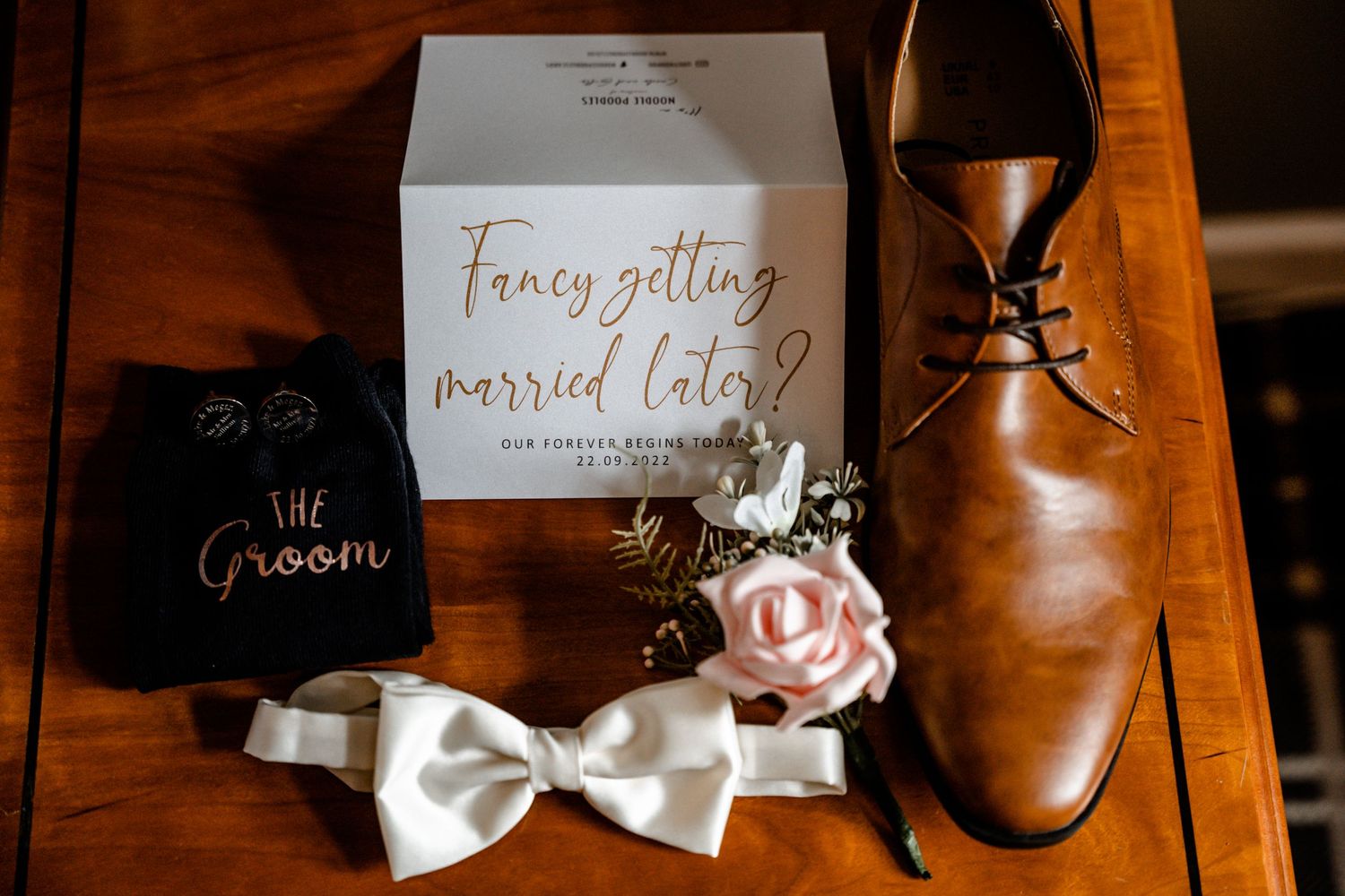 Wedding details with brown leather shoe, white bow tie, and elegant invitation on wooden surface.