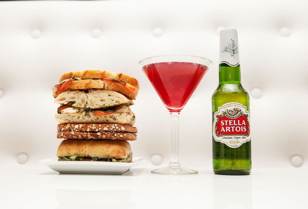 Gourmet burger stack with red cocktail and Stella Artois beer bottle on white background.