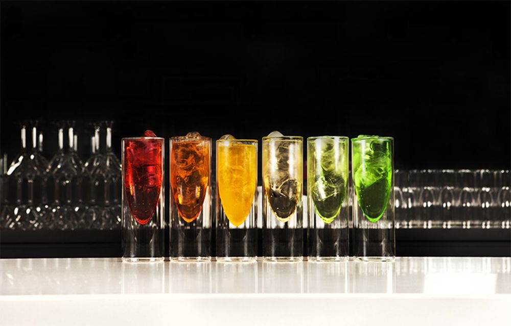 Rainbow colored drinks lined up in tall glasses on black background with dramatic lighting.