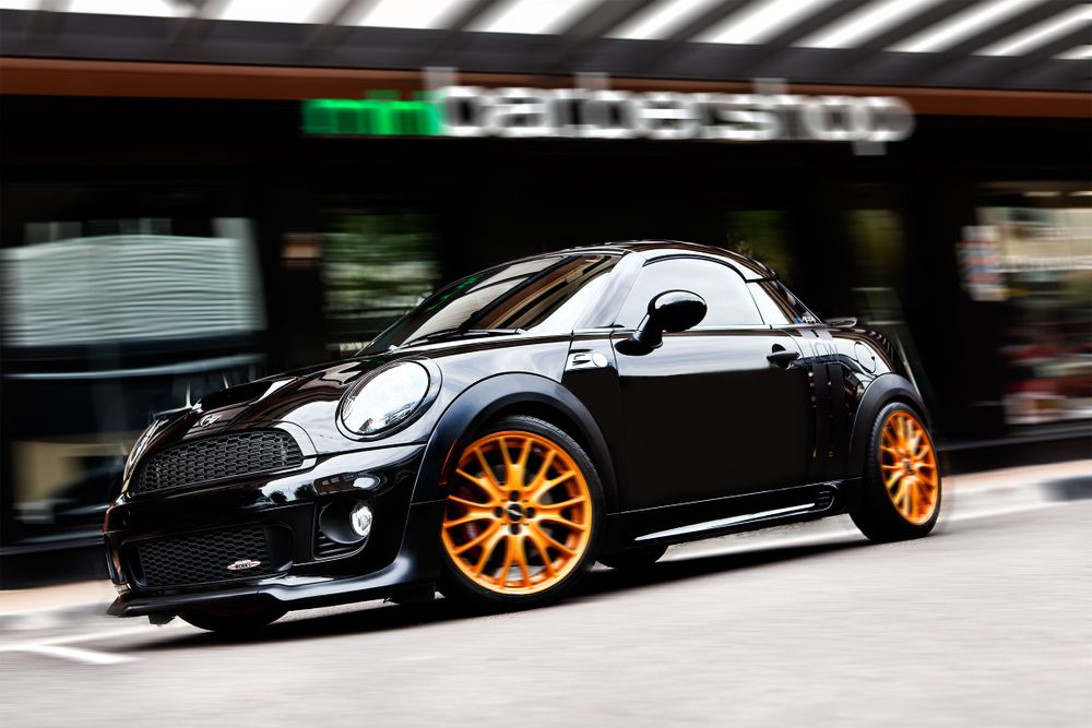Black Mini Cooper with custom orange wheels parked in front of a store.