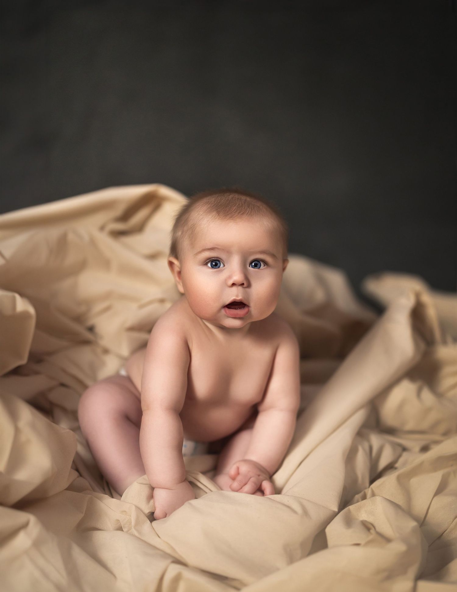 Baby sitting on soft beige fabric, gazing curiously. Toronto baby photography.