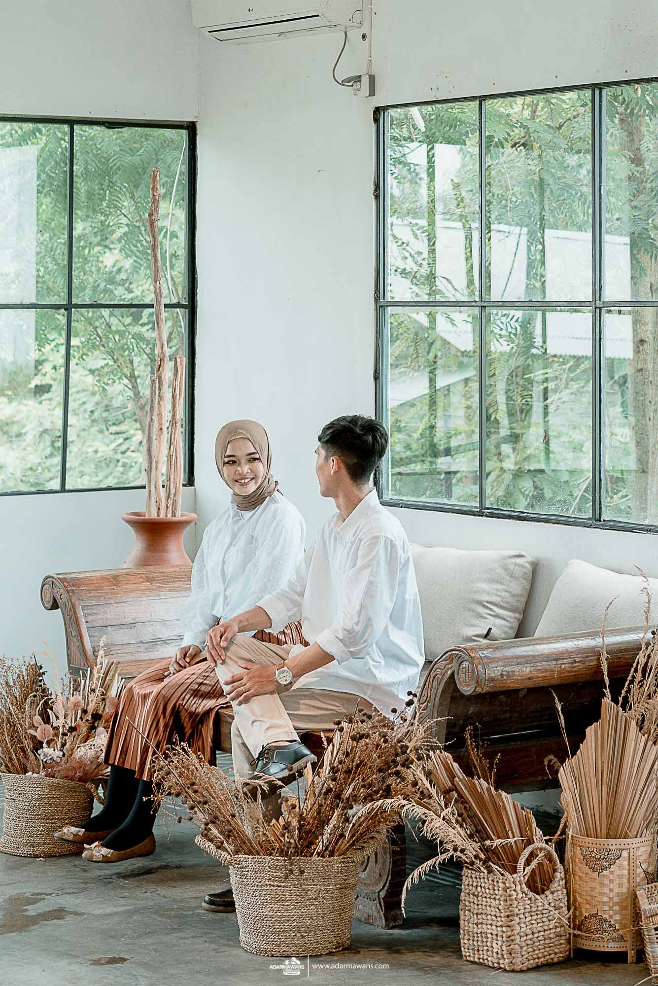 A couple in white outfits sits on a sofa surrounded by dried pampas grass and rattan baskets.
