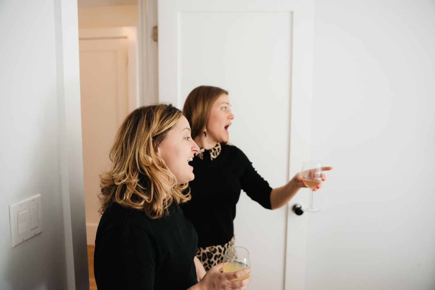 Two women laughing and pointing in candid moment against white wall.