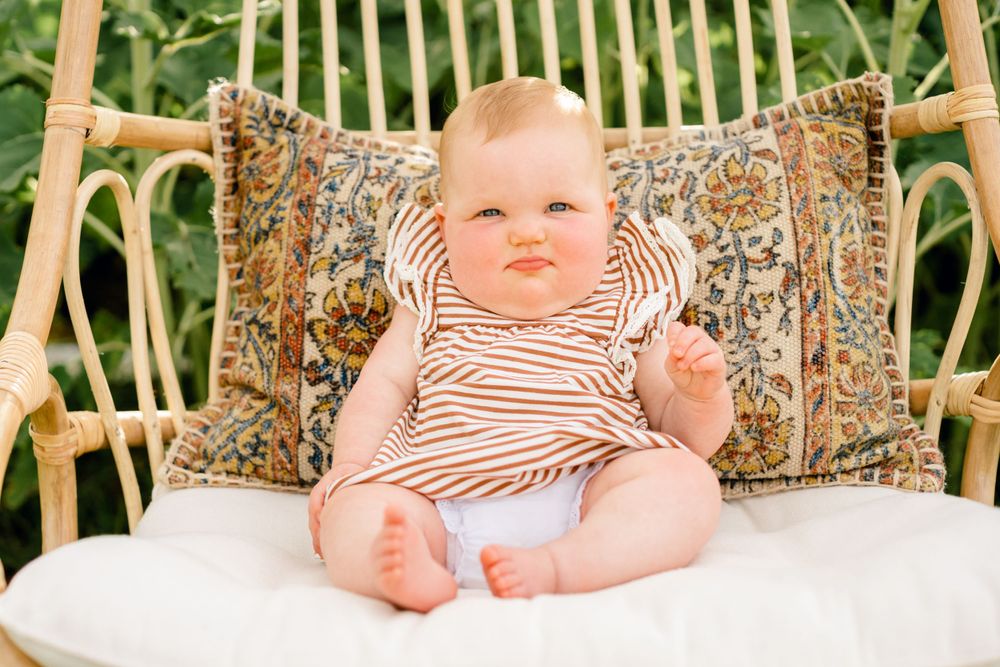 Baby in striped outfit sits on wicker chair with decorative pillows.
