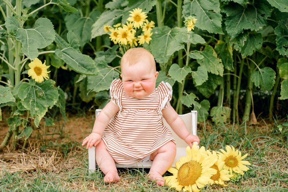 Baby in striped romper sits among sunflowers in garden setting.