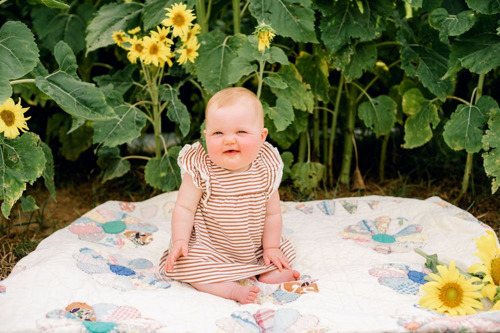 Baby in striped outfit sits on floral blanket surrounded by sunflowers.