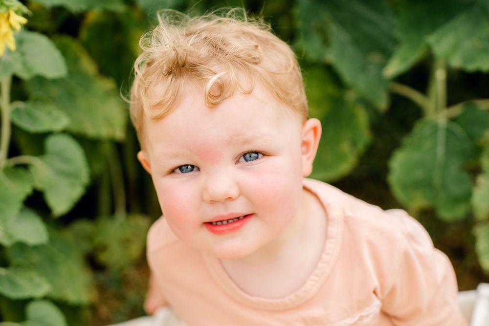 Toddler smiles brightly in peach colored shirt against sunflower garden backdrop.