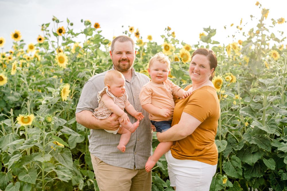 Family laughs together while posing for portrait in sunflower field during golden hour.