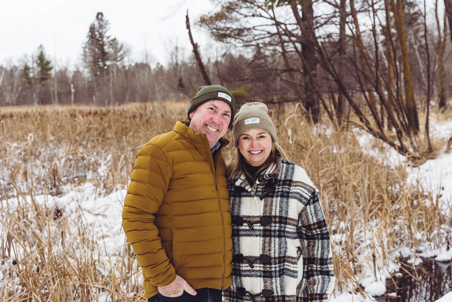 Couple wearing winter coats poses together in snowy field with brown grass and trees.