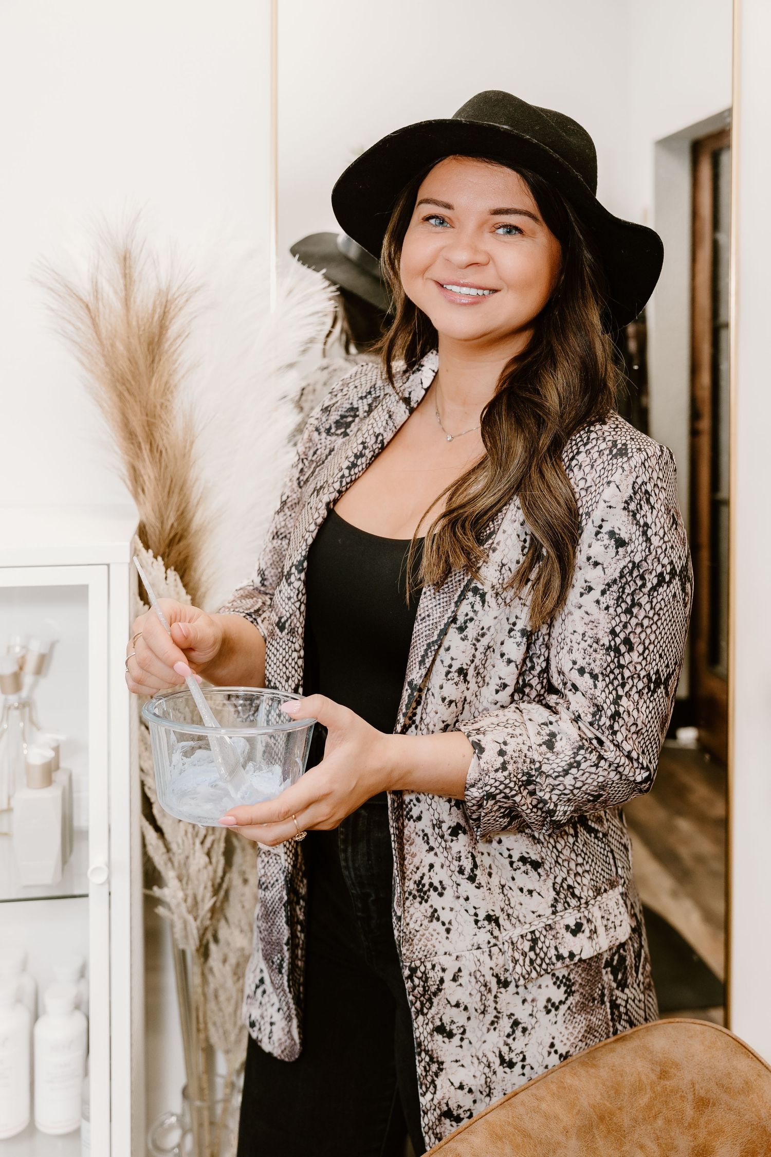 Hairstylist wearing black hat and snake print blazer holds pampas grass decor in stylish salon setting.