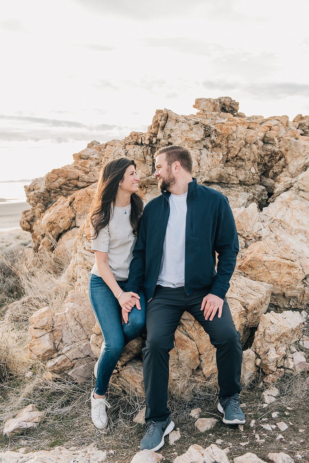 Couple sitting on rocky outcrop by ocean in casual attire.