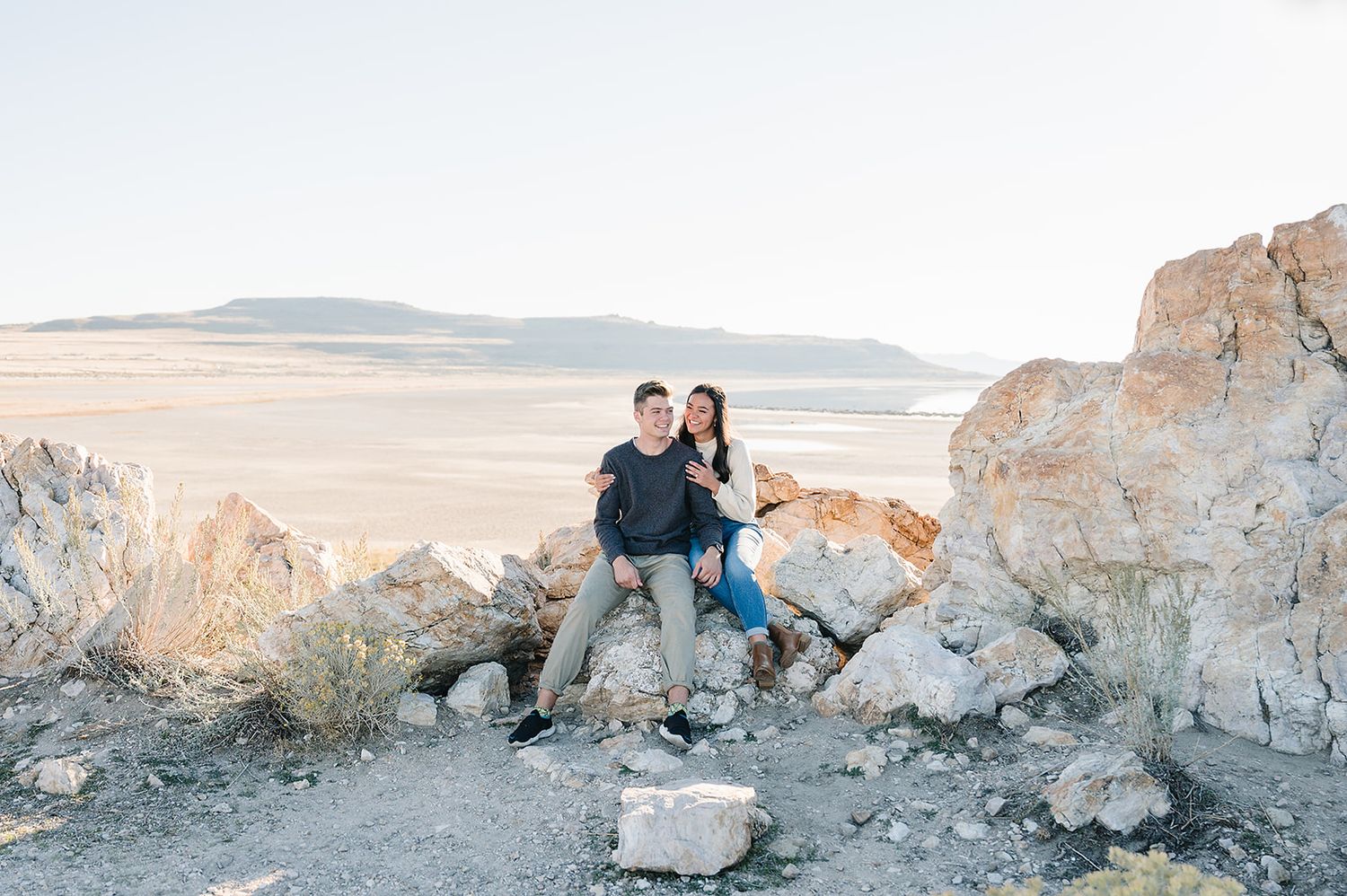 A couple sits on rocky terrain overlooking a desert landscape during sunset.