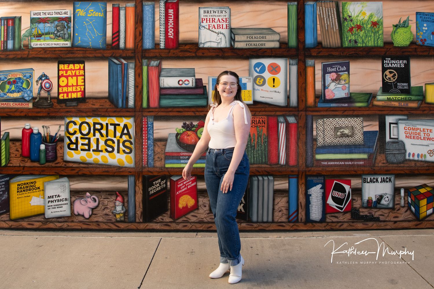 Portrait in front of colorful bookshelves showing 'Corita Kelsis' text and various book spines.