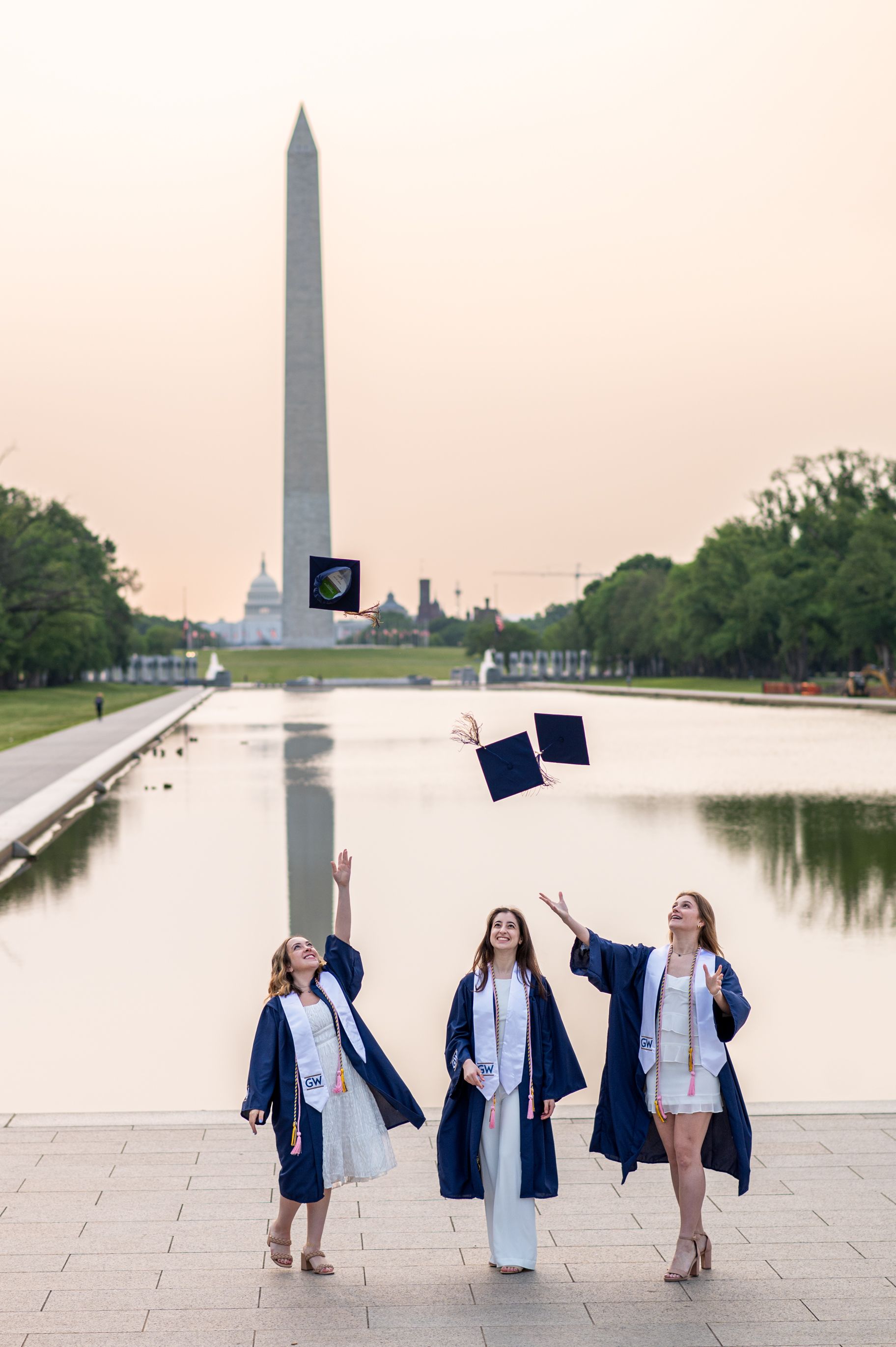 Three George Washington University graduates toss their caps in the air in front of the Lincoln Memorial reflecting pool