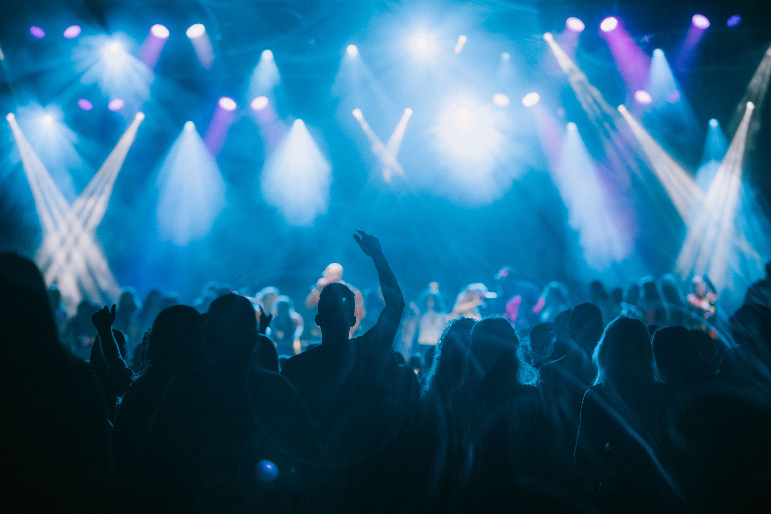 Concert crowd silhouetted against blue stage lighting with dramatic crossing spotlights.