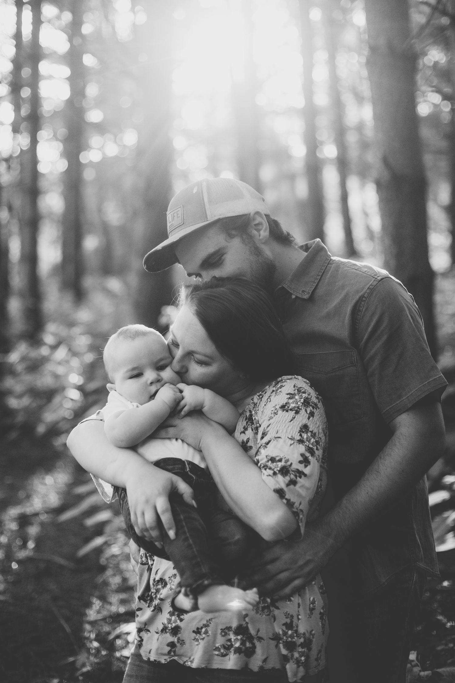 black & white photo of a mom and dad hugging their baby together; mom kisses the baby's cheek