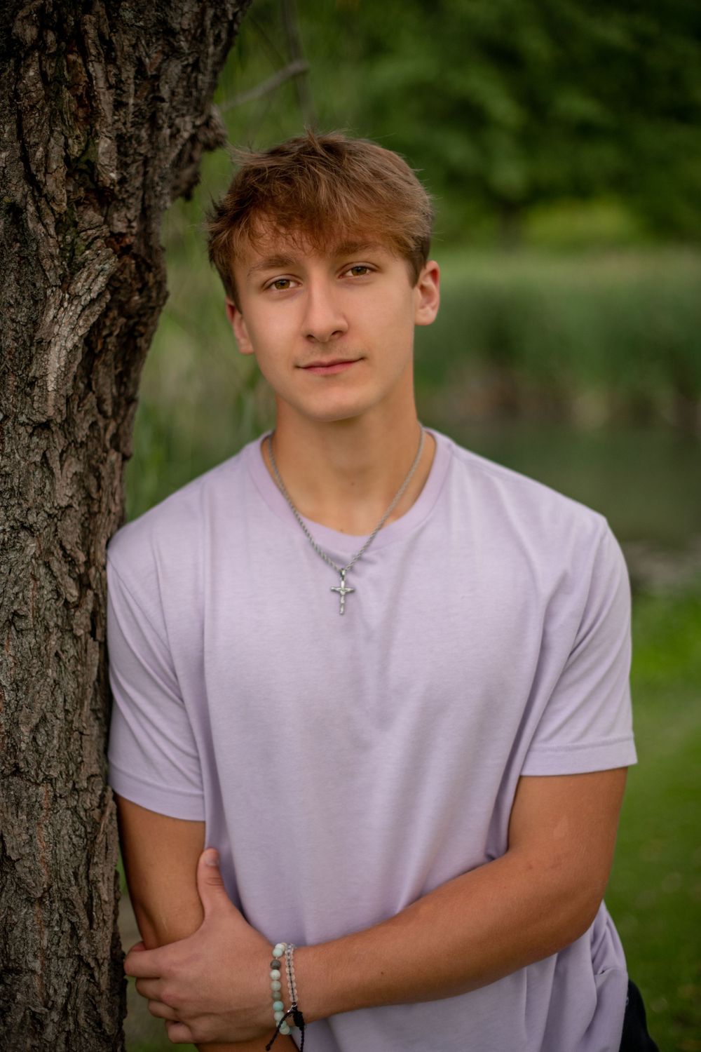 Portrait against tree trunk wearing lavender t-shirt with cross necklace.