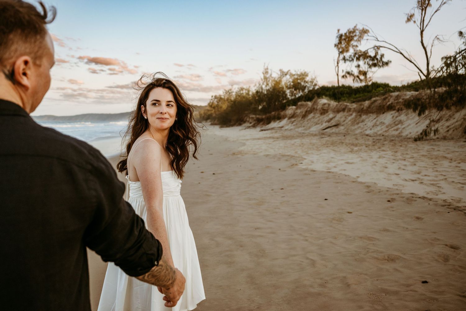 A romantic couple holding hands on a beach at sunset with wind-swept waves in the background.