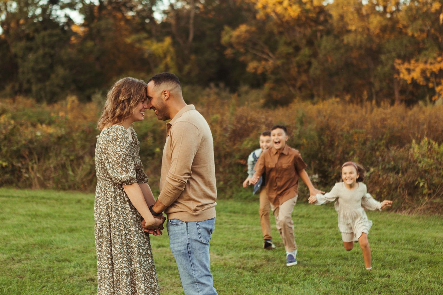 Couple shares romantic moment while children play in autumn field behind them.