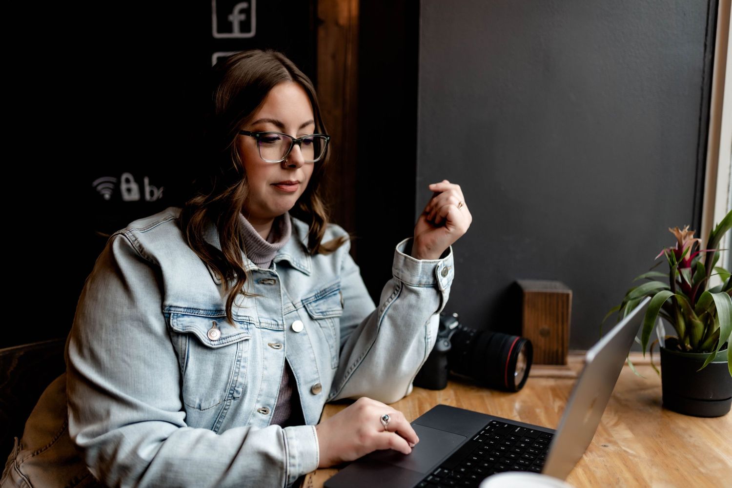 Woman in glasses and denim jacket works on laptop at wooden cafe table with plant nearby.