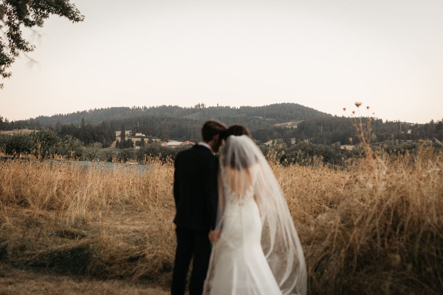 Two figures stand together in a field overlooking mountains at sunset.