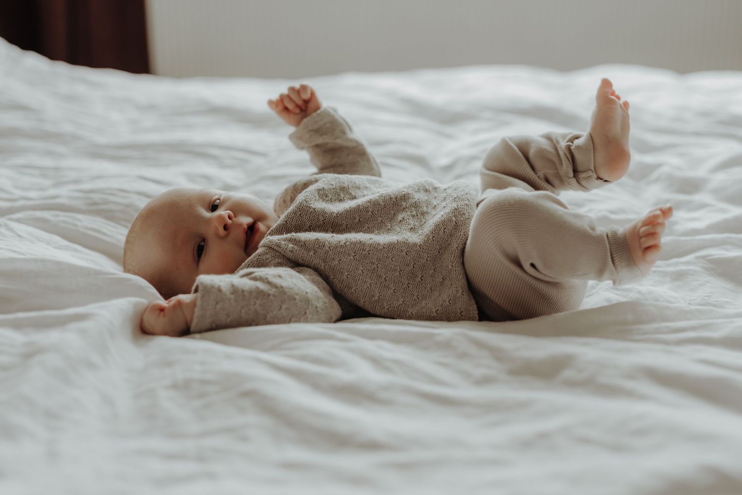 Baby wearing beige outfit lying on white bedding with arms outstretched.