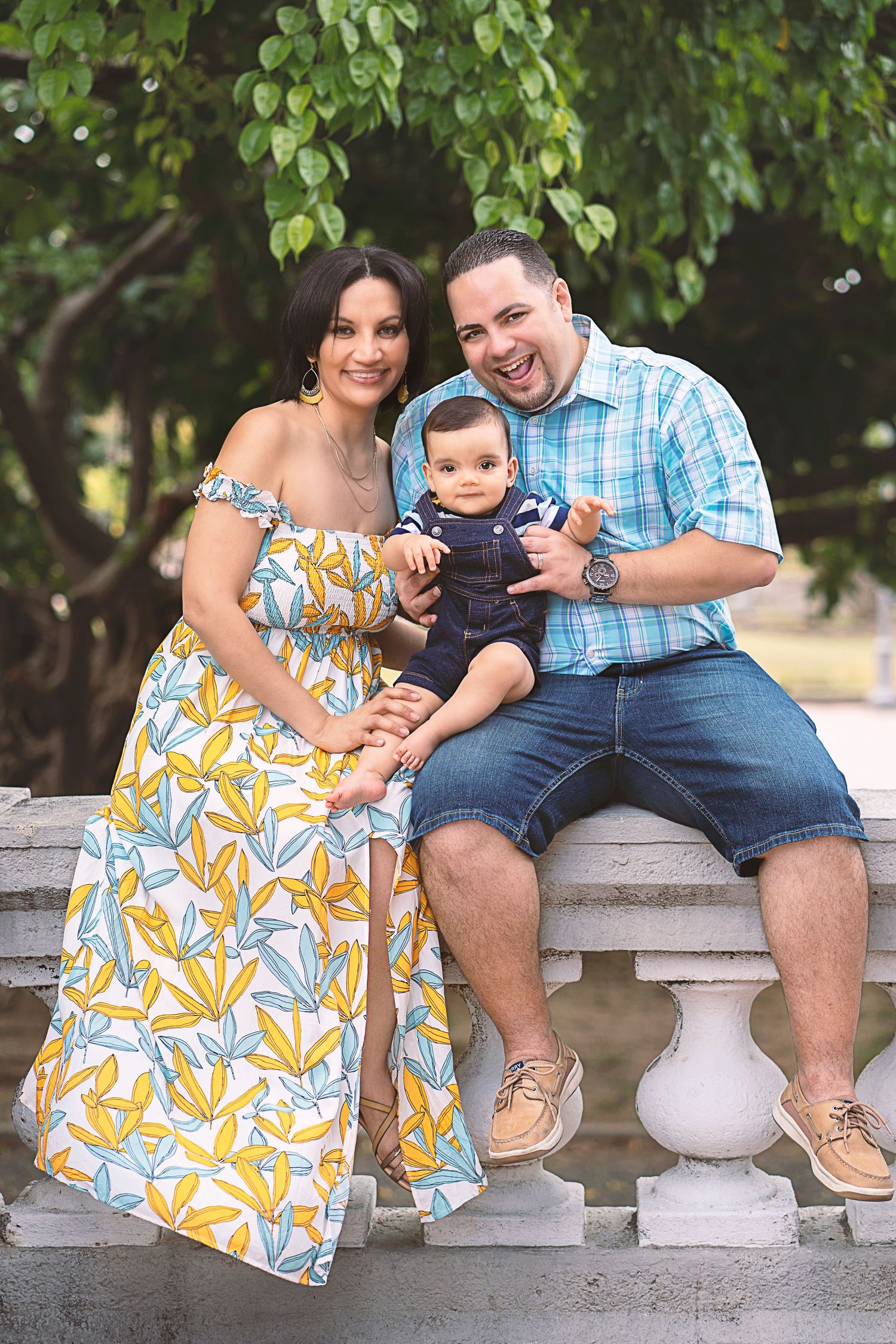 Family in summer attire sitting on stone railing under green trees.