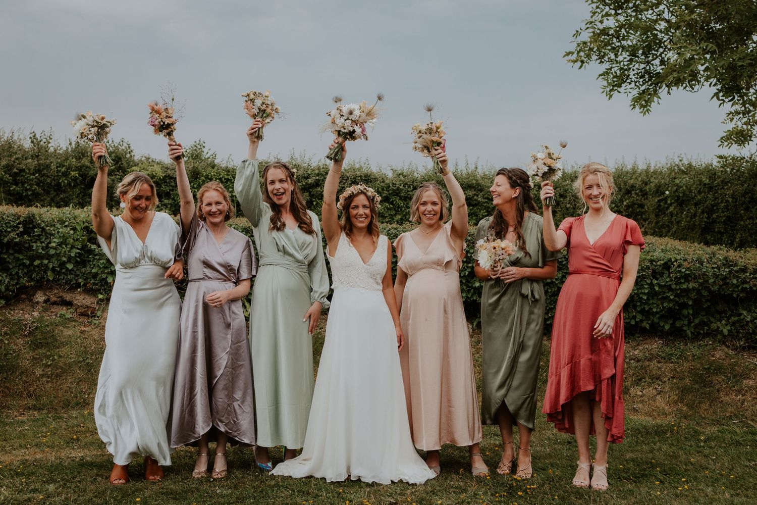 Wedding party in pastel dresses holding bouquets while celebrating outdoors.