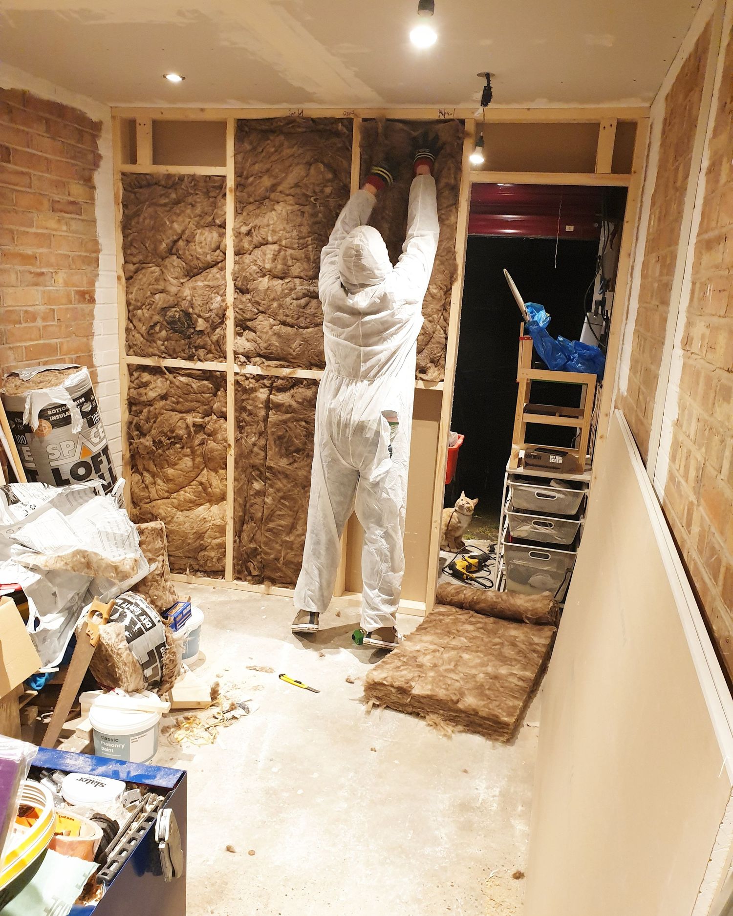Construction work in progress showing insulation installation between wall studs in brick-walled room.