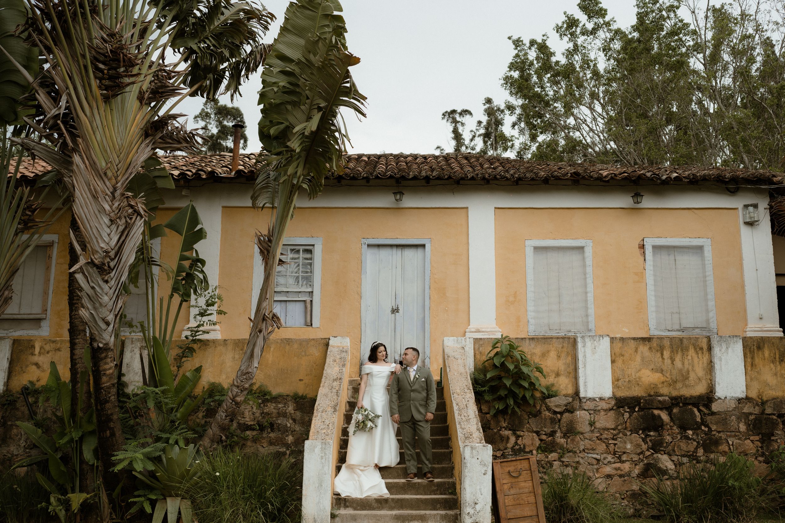 A couple in wedding attire pose together against a peach-colored colonial style building with palm trees.