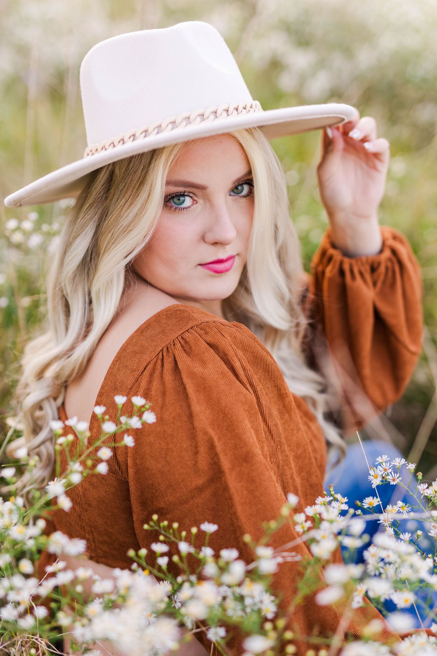 Portrait in wildflower field with cream hat and rust-colored dress among blue and white blooms.