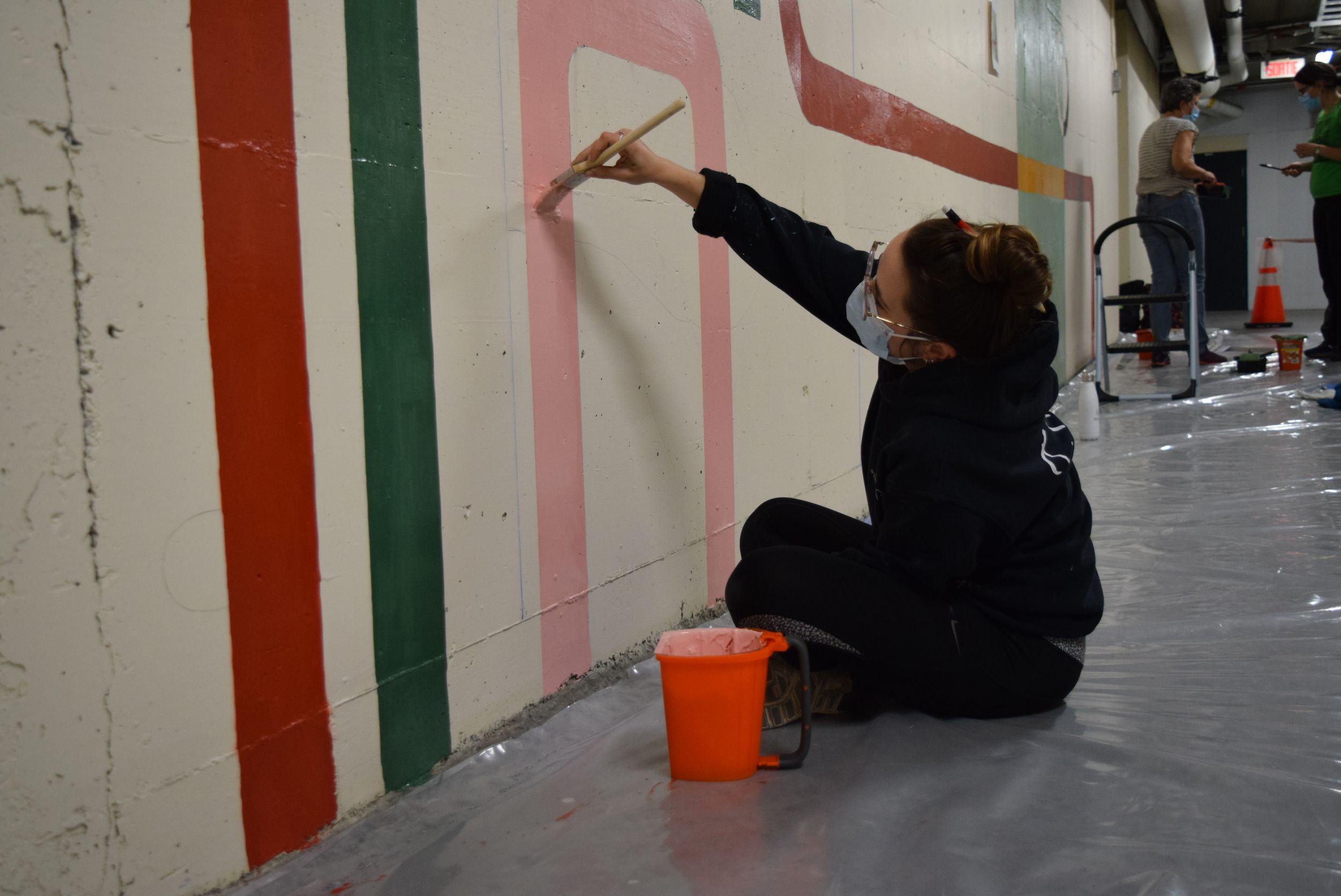 Artist sitting on floor painting geometric stripes in red, green, and pink on a wall mural.