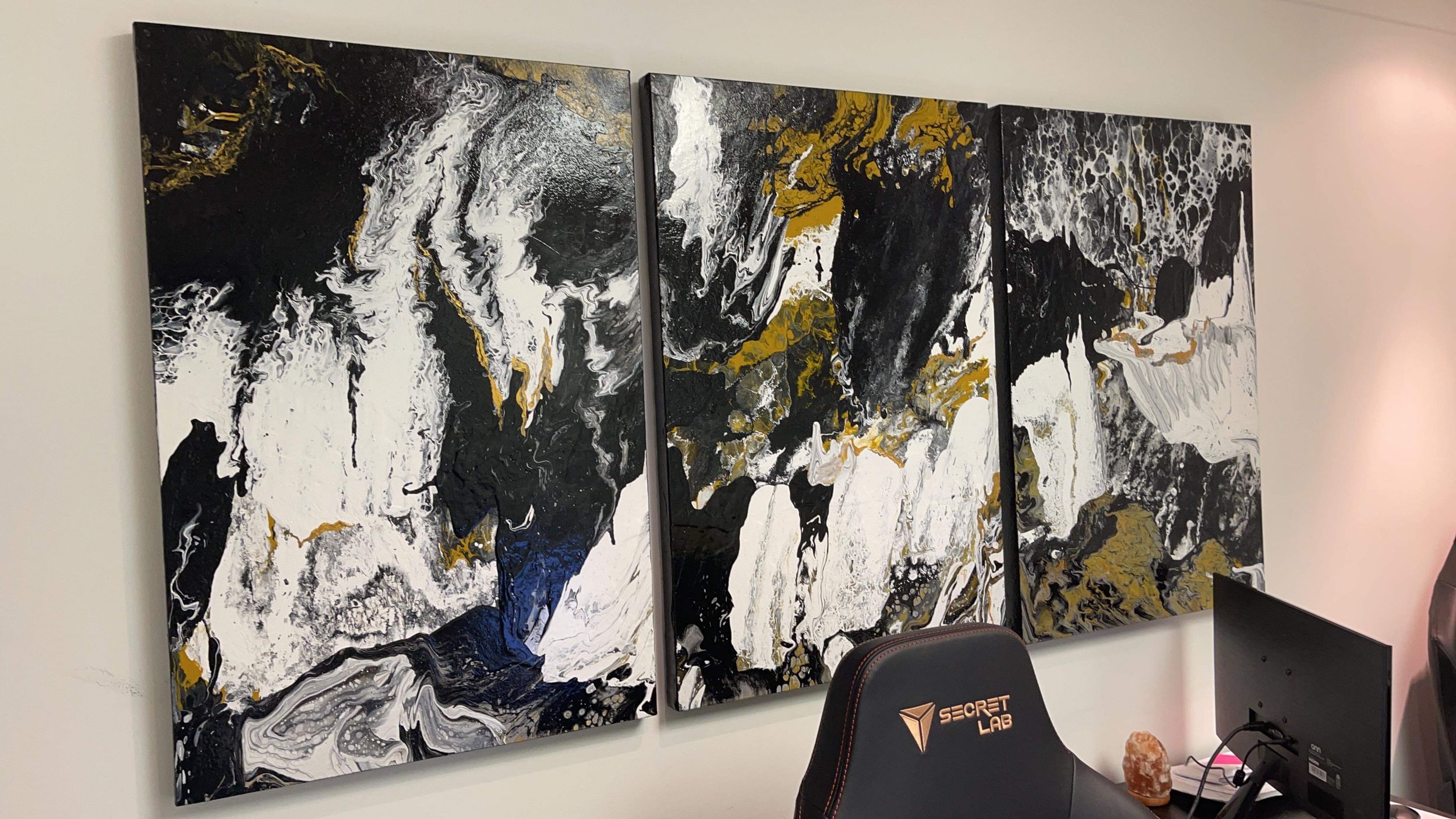 Abstract triptych canvas painting in black, white, gold, and blue displayed on an office wall.