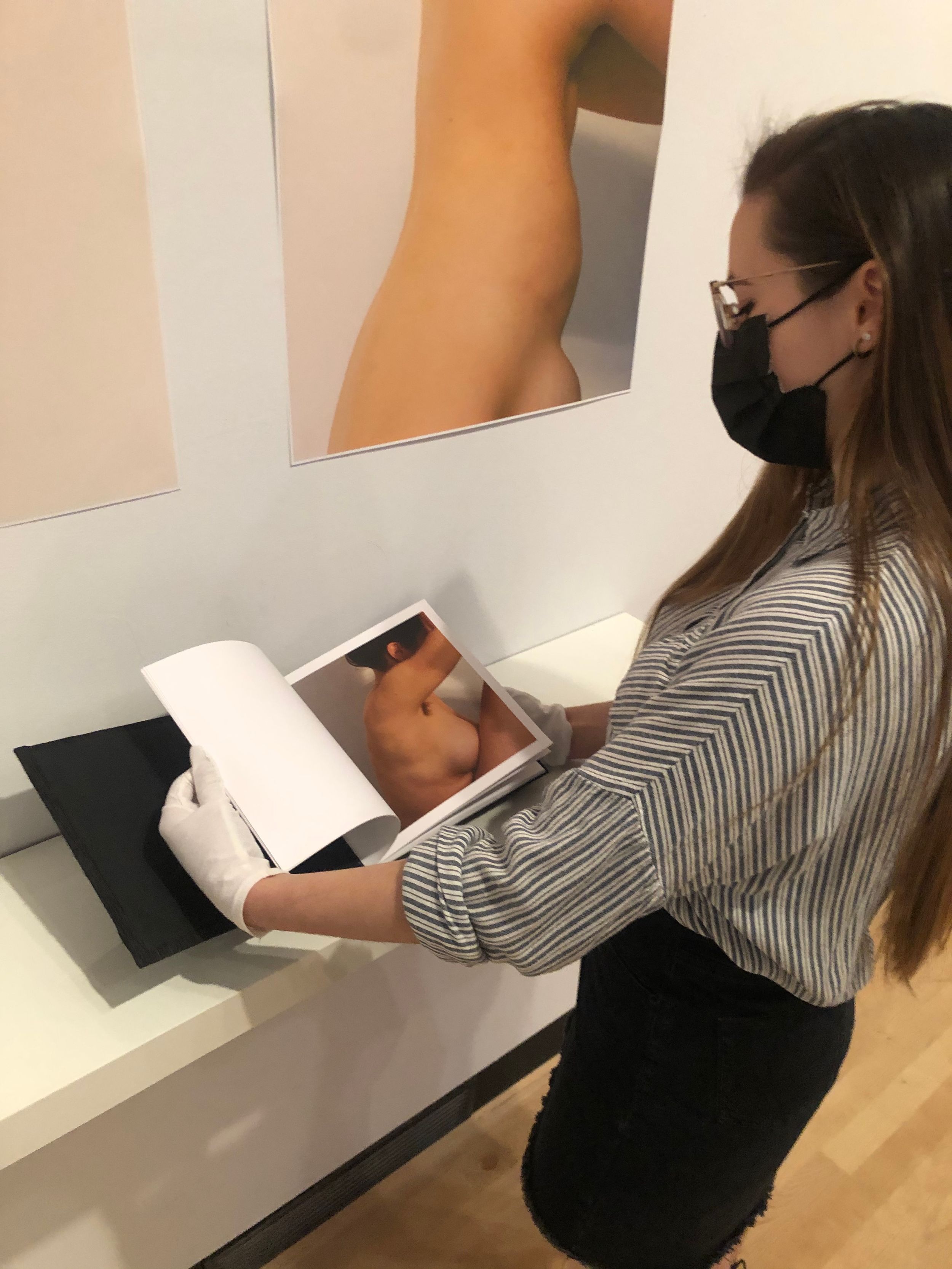 Masked woman in white gloves reviewing an art photography book beside wall-mounted prints in a gallery.